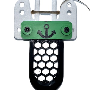 May include: A 3D-printed nautical-themed object featuring a white top section with holes, a green center piece with a black anchor, and a black honeycomb-patterned bottom. A brown string is attached.