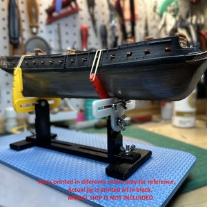 May include: A black model ship is held by a black, yellow, red, and white jig. The ship is detailed with dark wood and gold accents. The jig is on a blue surface. Text on the image states, "Parts printed in different colors only for reference. Actual jig is printed all in black. MODEL SHIP IS NOT INCLUDED."