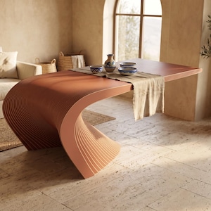 Puede incluir: Una mesa de comedor de color terracota que presenta una base curva continua con un diseño acanalado y en capas. El tablero es plano y rectangular, cubierto con un camino de mesa de tela clara y tres cuencos de cerámica estampados.