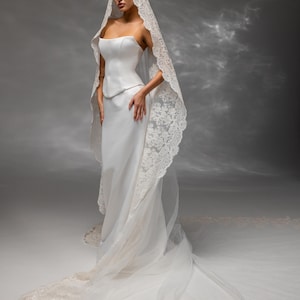 May include: A strapless white wedding gown with a long train. The dress features a fitted bodice and a flowing skirt. A lace-trimmed veil drapes over the head and shoulders, extending into a long train. The veil and train are adorned with delicate embellishments.