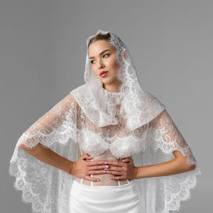 May include: A white lace bridal cape with a scalloped edge, paired with a matching white dress. The cape features delicate floral lace and a high neckline, creating an elegant and romantic look. The dress has a satin skirt.