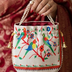 May include: A white drawstring bag with a colorful parrot design. The bag features a pearl beaded trim and handles, with red and gold accents. The parrots are red, blue, green, and yellow, perched on a branch with flowers.