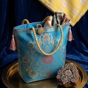 Könnte beinhalten: Eine türkisfarbene Tragetasche mit goldener Blumenstickerei, goldenen Seilgriffen und rosa Quasten. Die Tasche steht auf einem goldenen Tablett mit einem Holzblock mit floralem Design. Im Hintergrund befindet sich ein gelbes kariertes Tuch.