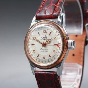 Puede incluir: Un reloj Oris vintage con caja plateada, bisel dorado y esfera color crema. El reloj presenta una correa de cuero marrón y muestra el texto "ORIS 17 JEWELS AUTOMATIC SWISS MADE".