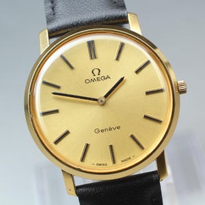 Pode incluir: Um relógio de pulso Omega Genève em tom dourado com mostrador redondo dourado, índices de bastão pretos e ponteiros pretos. O relógio possui uma pulseira de couro preta e exibe o logotipo Omega e o texto Genève, Swiss Made no mostrador.