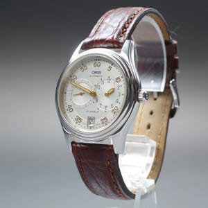 Peut inclure: Une montre-bracelet automatique Oris argentée avec un cadran crème et un bracelet en cuir marron. Le cadran de la montre présente des aiguilles et des chiffres dorés, avec un affichage de la date à 6 heures. Le bracelet a un motif crocodile.