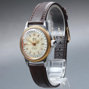Puede incluir: Reloj automático Oris vintage con esfera color crema, bisel dorado y correa de cuero marrón. El reloj presenta una visualización de la fecha y una caja plateada. El reloj se exhibe sobre un soporte transparente.