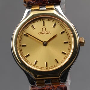 May include: A gold-toned Omega wristwatch with a gold dial and black hour markers. The watch has a gold bezel and a silver-toned case. The watch band is brown leather. The watch face displays the Omega logo.