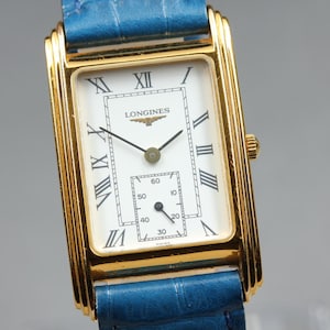 May include: A rectangular gold-tone wristwatch featuring a white dial with black Roman numerals, a sub-dial for seconds, and the Longines logo. The watch is attached to a textured blue leather strap with a visible grain pattern and a gold-tone crown.