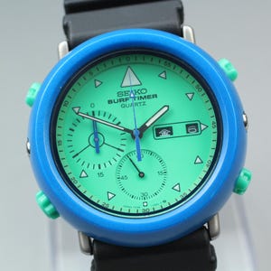 May include: A vintage Seiko Surf Timer quartz watch with a vibrant teal face and a blue bezel. The watch features a black rubber strap and green accents on the side buttons. The watch face displays multiple subdials and the text "SEIKO SURF TIMER QUARTZ".