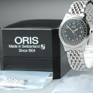 May include: A silver-tone Oris automatic wristwatch with a black dial, white Arabic numerals, and a date pointer hand. The watch features a multi-link metal bracelet and is displayed with its black presentation box and extra metal links.