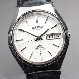 May include: A vintage Seiko automatic watch with a white face, black hands, and a date display. The watch has a silver-tone case and a black leather strap. The watch face includes the text "SEIKO AUTOMATIC LM Special 23 JEWELS".