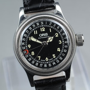 May include: An Oris Automatic wristwatch with a black dial, white Arabic numerals, and a date pointer hand. The watch features a silver-toned coin-edge bezel, a black leather strap, and a date track printed along the outer edge of the dial.