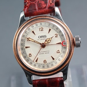 Könnte beinhalten: Eine Vintage-Armbanduhr von Oris mit cremefarbenem Zifferblatt, goldenen Zeigern und Datumsanzeige. Die Uhr hat ein Edelstahlgehäuse, eine goldfarbene Lünette und ein braunes Lederarmband. Das Zifferblatt zeigt "ORIS 17 JEWELS AUTOMATIC SWISS MADE".