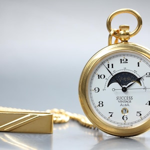 May include: A gold pocket watch with a white face and black numerals. The watch has a moon phase indicator and the text "SUCCESS VINTAGE ALBA". A matching gold chain and rectangular fob are also visible.