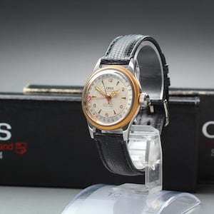 Puede incluir: Un reloj de pulsera automático Oris con esfera color crema, bisel dorado y correa de cuero negro texturizado. La esfera muestra números arábigos, una aguja de fecha y el logotipo de Oris. Se presenta junto a una caja negra de la marca Oris.
