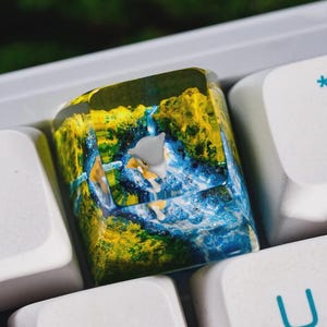 May include: A custom keycap featuring a miniature figurine set within a clear resin block. The keycap has a landscape design with yellow and blue colors, and is surrounded by white keyboard keys.