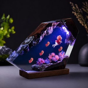 May include: A decorative lamp featuring a dark wood frame encasing a clear resin block. Inside, a blue-lit underwater scene displays a small figure in a red dress swimming among pink coral and several small orange fish figures on a wooden base.