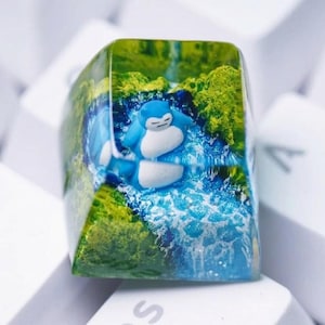 May include: A custom keyboard keycap with a blue and white cartoon character in a waterfall scene. The keycap is encased in clear resin, with green and blue accents. The keycap is set against a background of white keyboard keys.