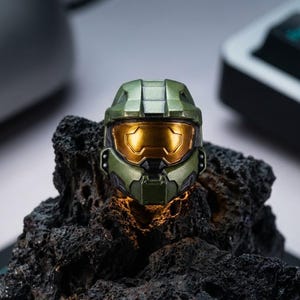 Puede incluir: Una tecla verde oliva con forma de casco de Master Chief, con una visera dorada. Descansa sobre una superficie oscura y texturizada. La tecla está iluminada desde dentro, creando un efecto brillante.