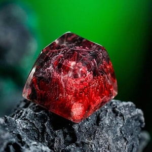 May include: A clear, geometric resin object with a vibrant red interior, resting on a dark, textured surface. The red design has a swirling, organic pattern. The background is a blurred green.