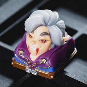 Może przedstawiać: Niestandardowa nasadka na klawisz przedstawiająca stylizowaną głowę wampira z białymi włosami, czerwonymi oczami i krwią. Nasadka ma fioletowy kołnierz, złote akcenty i niebieską podstawę na ciemnym tle.