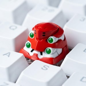 May include: A red and white custom mechanical keyboard keycap with green accents. The keycap has a robotic design and is set among white keyboard keys. The keycap is a unique accessory for computer enthusiasts.