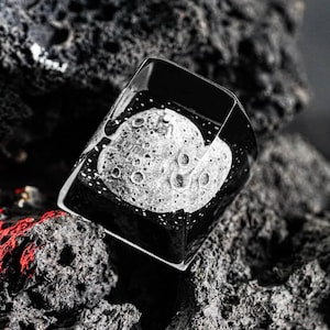 May include: A white ceramic mug with a black handle and the text "But First, Coffee" in a bold, black font. The mug is set against a blurred background, suggesting a cozy setting.