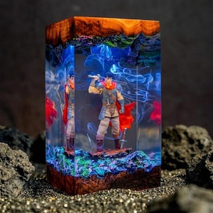 May include: A rectangular resin sculpture featuring a warrior figure with a sword, encased in a block with a wood top and a colorful, smoky interior. The figure wears a red cape and is set against a backdrop of blue and green hues.