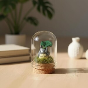 May include: A small gray creature figurine with a cream-colored belly and dark markings, holding a green leaf umbrella, stands on a patch of artificial grass inside a clear glass dome with a light brown cork base, creating a miniature display.