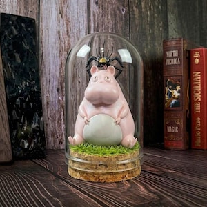 Puede incluir: Una figura de hipop&oacute;tamo rosa con vientre blanco sentada sobre un trozo de hierba verde dentro de una campana de cristal. Una figura de ara&ntilde;a negra est&aacute; posada encima. La base es de corcho. Art&iacute;culo decorativo.