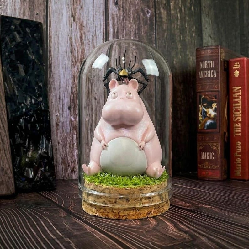 Puede incluir: Una figura de hipop&oacute;tamo rosa con vientre blanco sentada sobre un trozo de hierba verde dentro de una campana de cristal. Una figura de ara&ntilde;a negra est&aacute; posada encima. La base es de corcho. Art&iacute;culo decorativo.
