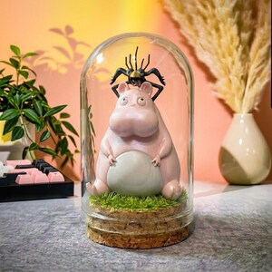 Puede incluir: Una c&uacute;pula de cristal decorativa con una figura de hipop&oacute;tamo rosa y un insecto negro. El hipop&oacute;tamo est&aacute; sobre una base de c&eacute;sped artificial y corcho. El fondo es de color melocot&oacute;n c&aacute;lido. Un objeto decorativo.