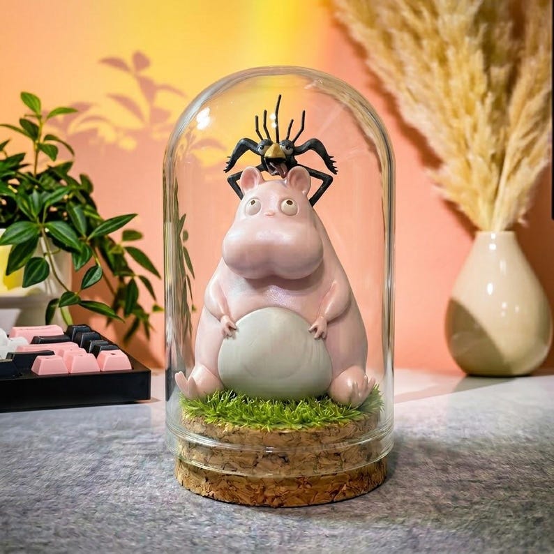 Puede incluir: Una c&uacute;pula de cristal decorativa con una figura de hipop&oacute;tamo rosa y un insecto negro. El hipop&oacute;tamo est&aacute; sobre una base de c&eacute;sped artificial y corcho. El fondo es de color melocot&oacute;n c&aacute;lido. Un objeto decorativo.