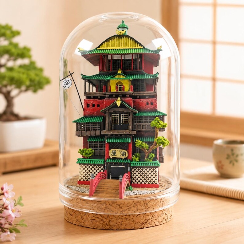 K&ouml;nnte beinhalten: Ein Miniaturmodell eines mehrst&ouml;ckigen Geb&auml;udes mit roten, gr&uuml;nen und gelben architektonischen Details, eingeschlossen in einer klaren Glaskuppel auf einem Korksockel. Das Modell zeigt eine kleine rote Br&uuml;cke, eine wei&szlig;e Flagge und ein Schild mit dem Text 油.