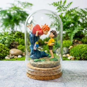 May include: A glass cloche containing a figurine of a small orange-haired character in a red dress leaning over a blue wave, facing a character with brown hair in a yellow shirt and brown shorts holding a green bucket on a rocky base.