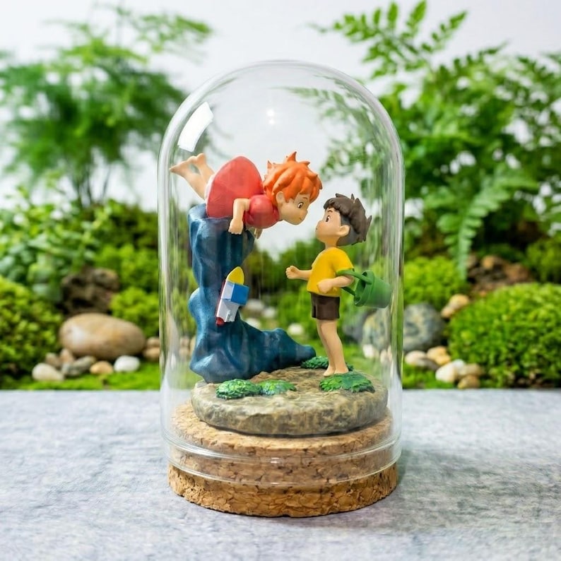 May include: A glass cloche containing a figurine of a small orange-haired character in a red dress leaning over a blue wave, facing a character with brown hair in a yellow shirt and brown shorts holding a green bucket on a rocky base.
