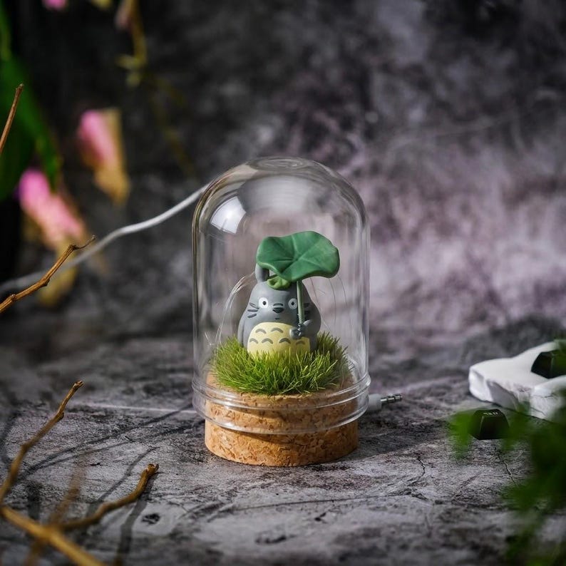 May include: A small gray and cream-colored creature figurine with a green leaf umbrella, standing on a patch of artificial green grass inside a clear glass dome with a cork base. The creature has a rounded body, whiskers, and a patterned belly.
