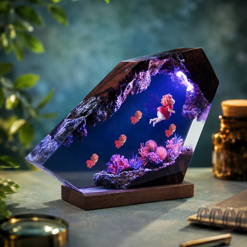 May include: A decorative lamp featuring a dark wood frame enclosing a clear resin block. Inside the resin, a miniature scene depicts a small figure in a red dress swimming among pink coral formations and several small orange fish, illuminated by blue light.