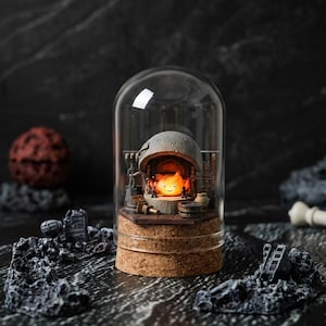 Puede incluir: Una cúpula de cristal decorativa que presenta una escena en miniatura con un fuego naranja brillante y una cara sonriente. La base es de corcho, y la escena incluye herramientas en miniatura y un horno de piedra. La cúpula está sobre un fondo oscuro.