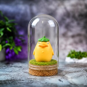 May include: A yellow figurine of a duck-like creature with a green hat, inside a glass dome. The figurine sits on a bed of green moss, atop a cork base. The dome is clear, and the background is blurred.