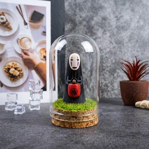 Op de afbeelding: Een figuurtje van een in het zwart gekleed personage met een wit masker dat een kleine rode lantaarn met zwarte Japanse tekst vasthoudt, in een glazen stolp op een kurken voet met een plukje kunstmatig groen gras erin.
