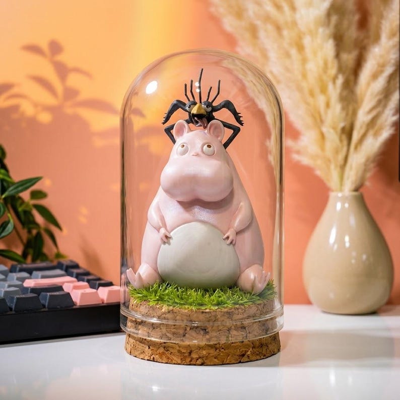 Puede incluir: Una c&uacute;pula de cristal decorativa caprichosa con una figura de hipop&oacute;tamo rosa con una ara&ntilde;a negra encima. El hipop&oacute;tamo est&aacute; sentado sobre un parche de hierba verde, sobre una base de corcho. La c&uacute;pula se exhibe sobre una superficie blanca.