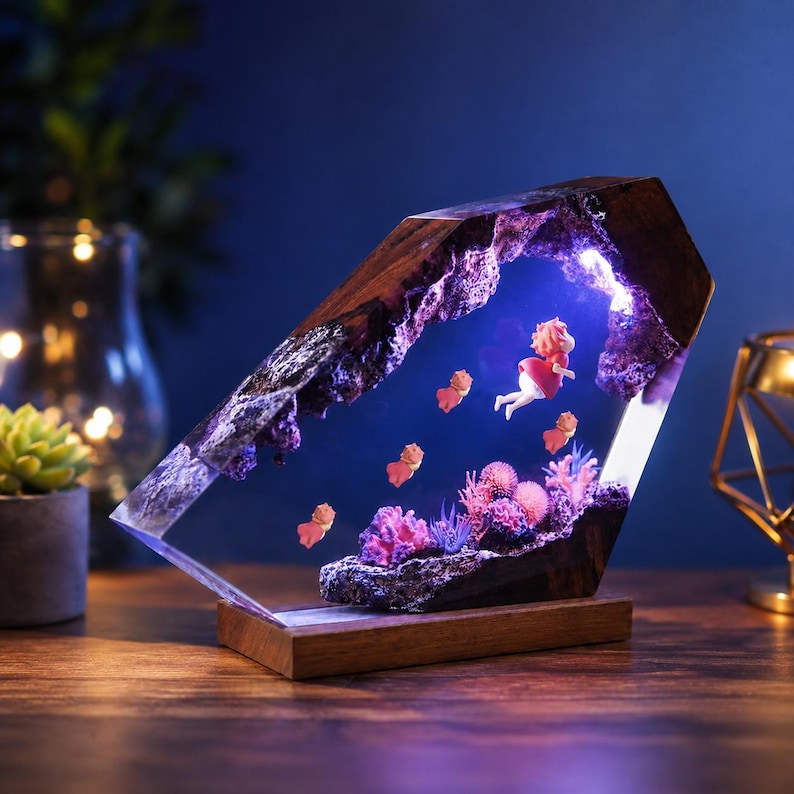 May include: A decorative lamp featuring a dark wood frame with a clear resin centre. Inside the resin, a small figure in a red dress swims among miniature pink and purple coral structures, illuminated by internal purple and white LED lighting.