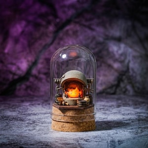 Pu&ograve; includere: Un diorama in miniatura racchiuso in una cupola di vetro trasparente su base di sughero, raffigurante un piccolo focolare in pietra con all'interno un personaggio a forma di fiamma arancione brillante, circondato da minuscoli attrezzi e barili.