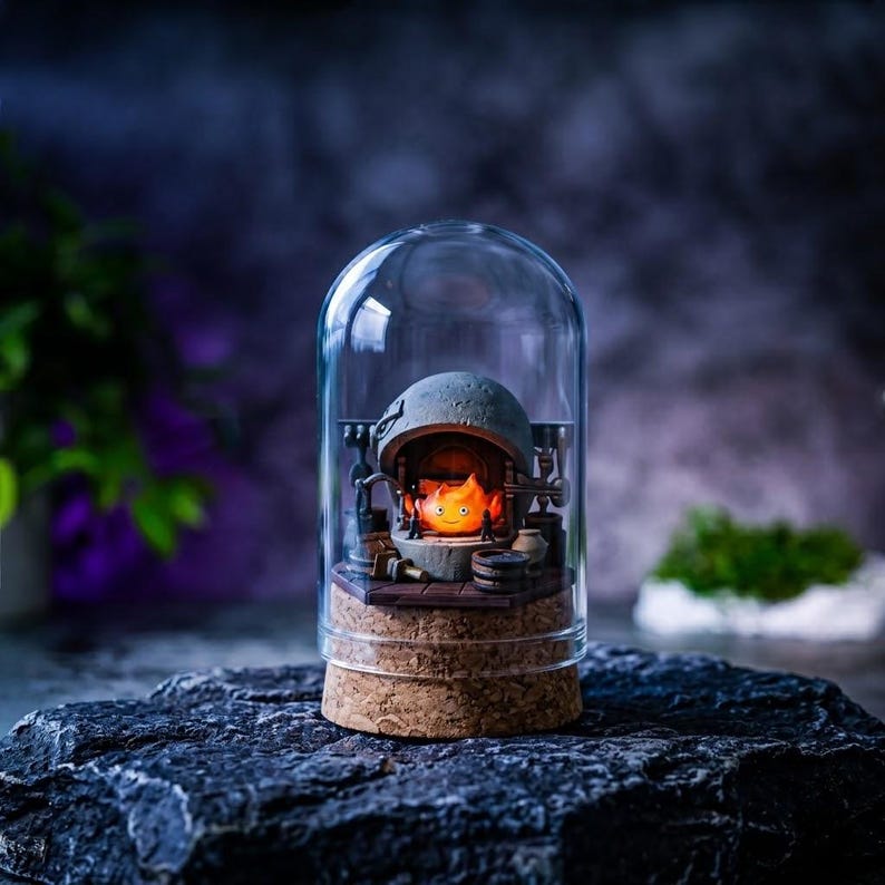 Pu&ograve; includere: Una cupola in vetro trasparente con base in sughero contenente un focolare in pietra grigia in miniatura. All'interno del focolare si trova un piccolo personaggio a forma di fiamma arancione brillante con un volto sorridente.