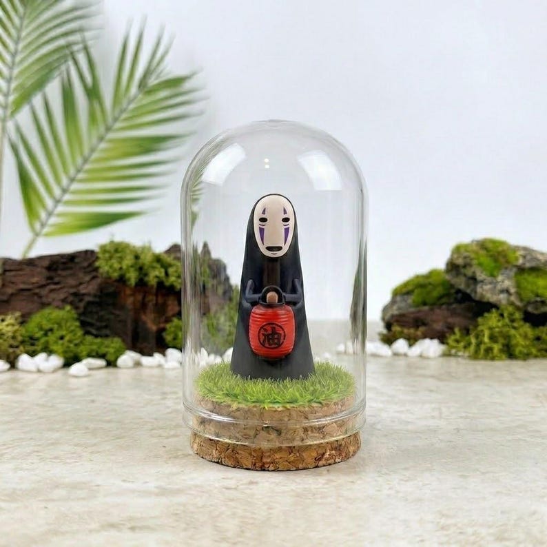 Op de afbeelding: Een figuurtje van een zwart, spookachtig personage met een wit masker en paarse markeringen, dat een kleine rode lantaarn met zwarte Japanse tekst vasthoudt. Het figuurtje staat op groen gras in een glazen stolp met een kurken voet.