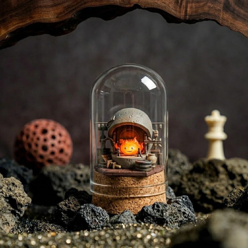 Pu&ograve; includere: Un diorama in miniatura racchiuso in una cupola di vetro con base in sughero, raffigurante un piccolo personaggio a forma di fiamma arancione brillante all'interno di una struttura a focolare color pietra con minuscoli accessori in legno.