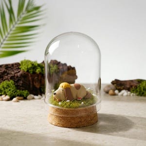 May include: A glass cloche with a cork base houses a sleeping dog figurine. The dog is light brown with a yellow and brown head, resting on a bed of green grass and tiny white flowers. The cloche sits on a light-colored surface.