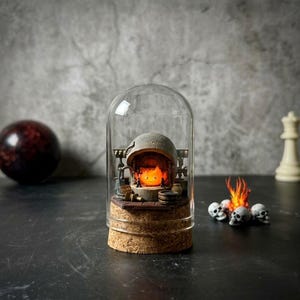 Pu&ograve; includere: Una cupola di vetro con base in sughero contenente un focolare in pietra grigia in miniatura con un personaggio a forma di fiamma arancione brillante all'interno. Accanto alla cupola si trova un piccolo mucchio decorativo di teschi bianchi.